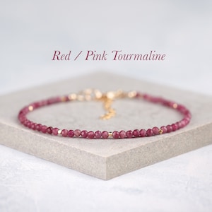 May include: A delicate bracelet featuring small, faceted red and pink tourmaline beads interspersed with tiny gold-toned metal spacers. The jewellery includes a gold-toned metal clasp and an extender chain. The text Red / Pink Tourmaline is visible.