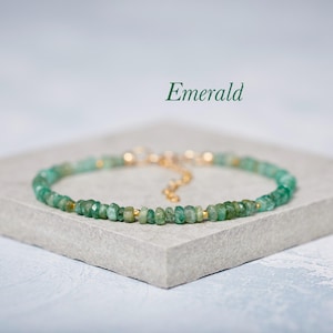 May include: An emerald bracelet featuring small, irregularly shaped green beads linked by a gold-coloured chain and accents. The word "Emerald" is written in green script. The bracelet is displayed on a grey surface.