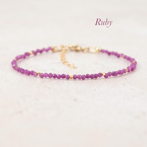 May include: A delicate bracelet featuring small, faceted, deep pink ruby beads interspersed with tiny gold-toned metal spacers. The piece includes a gold-toned metal chain extender and a clasp. The word Ruby is printed in the upper right corner.
