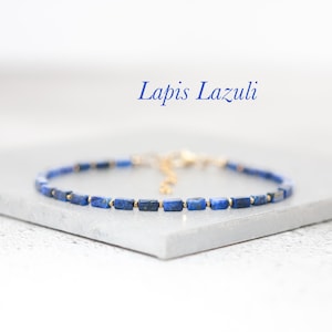 May include: A delicate bracelet made with small, rectangular lapis lazuli beads and gold spacers. The bracelet is on a white surface and the text "Lapis Lazuli" is written above it.