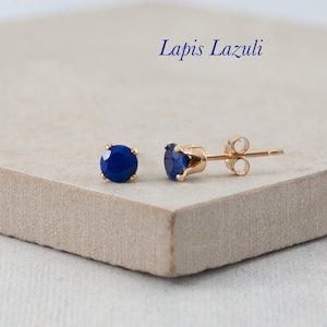 May include: Gold stud earrings featuring round, deep blue lapis lazuli gemstones. The earrings are set in a four-prong setting and are presented on a neutral surface. The text "Lapis Lazuli" is written in a dark blue script.
