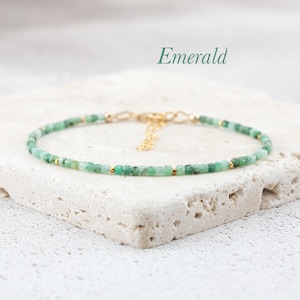 May include: A delicate gold chain bracelet with small emerald green beads. The bracelet is on a white marble surface. The word "Emerald" is written in black text above the bracelet.