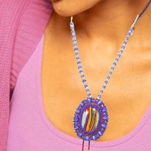 May include: A beaded necklace with a large, oval pendant. The pendant has a central design of colourful stripes within a blue beaded frame. The necklace has a blue beaded chain and a decorative tassel. The text "8124 Laleh" is visible.