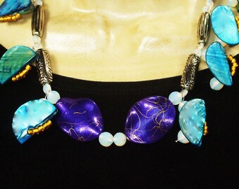 May include: A necklace featuring semi-circular blue shell segments, two central oval purple beads with gold-toned veining, silver-toned cylindrical spacers, and small white round beads, all arranged on a string against a black background.