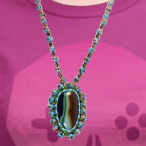 May include: A beaded necklace with a large oval pendant. The pendant features a dark, striped stone surrounded by turquoise and green beads. The necklace is composed of smaller turquoise and green beads. The background is a solid maroon color.
