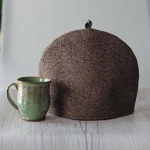 May include: A green ceramic mug with a textured design and a brown tea cosy with a leaf pattern. The cosy is dome-shaped with a small loop at the top. The mug is next to the tea cosy.