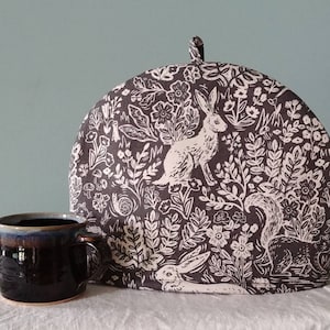 May include: A brown tea cozy with a white floral and rabbit print. The cozy has a round shape and a pointed top.