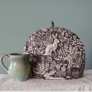 Könnte beinhalten: Ein brauner und weißer Teekosename mit einem Waldmuster mit Hasen und Blumen. Der Kosename steht auf einer weißen Oberfläche neben einer grünen Keramiktasse.