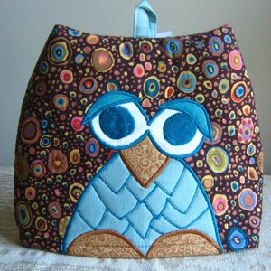 May include: A brown tea cozy with a colorful pattern of circles and an embroidered blue owl with white eyes.