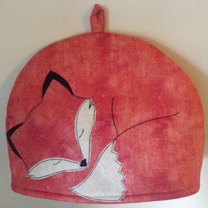 May include: Red tea cozy with a black and white fox design. The fox is curled up and sleeping.
