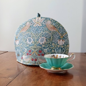 Könnte beinhalten: Eine teelafarbene Teekanne mit einem floralen Muster mit Vögeln und Erdbeeren. Die Teekanne steht auf einem Holztisch neben einer teelafarbene Teetasse mit einem floralen Muster und goldenen Verzierungen.