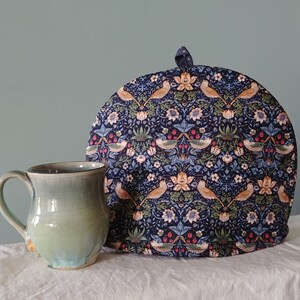 May include: A blue and white tea cozy with a floral pattern and birds. The cozy is sitting on a white surface next to a green ceramic mug.
