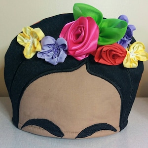 Puede incluir: Un calienta-tetera de tela negro y marrón con forma de cabeza de mujer con una corona de flores de colores. Las flores están hechas de tela e incluyen rosa, amarillo, rojo, verde y morado.