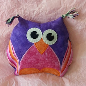 May include: Purple and pink owl-shaped pillow with orange beak and large white eyes. The pillow has a striped pattern on the sides and a floral pattern on the front.
