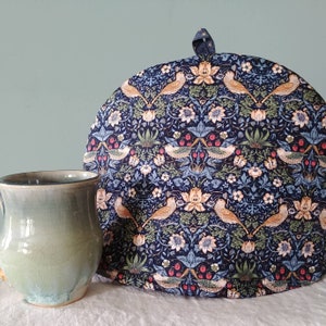 May include: A blue and white tea cosy with a floral pattern featuring birds. The cosy is round and has a scalloped edge.