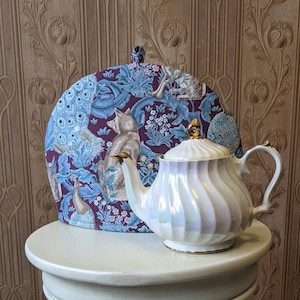 May include: A white teapot with a gold lid and handle, sitting on a white table. The teapot is decorated with a swirling pattern. A fabric tea cosy with a peacock and fox design is behind the teapot.