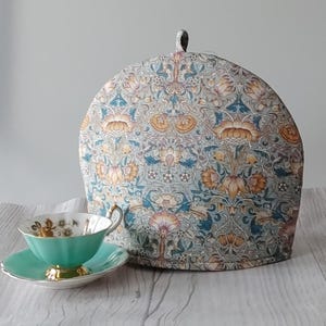 May include: A teal blue tea cozy with a floral pattern in shades of orange, yellow, and green. The cozy is made of fabric and has a dome shape.