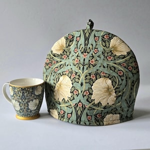 May include: A tea set featuring a floral patterned tea cozy and matching mug. The cozy is dome-shaped with a black handle, and the mug has a gold rim and base. The pattern includes cream-colored flowers, green leaves, and small pink blossoms.