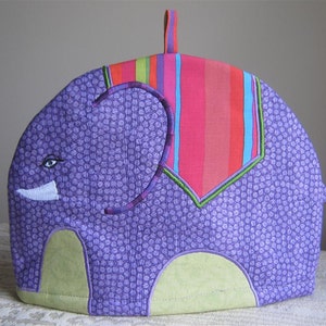 May include: Purple and green tea cozy shaped like an elephant with a striped pink, red, and green design on the back.