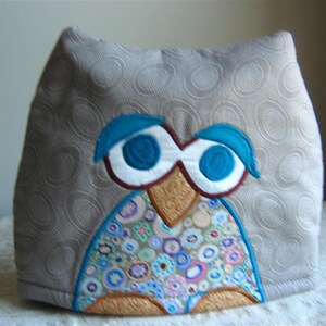 May include: A gray fabric teapot cozy with an embroidered owl design. The owl has large blue eyes, a brown beak, and a colorful patterned body.