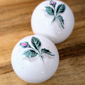 Puede incluir: Dos esferas de cerámica blanca con un diseño floral. Cada esfera presenta un solo capullo de rosa rosa con hojas verdes.