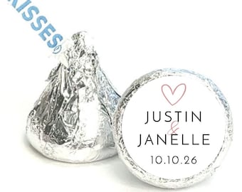 May include: Two foil-wrapped chocolate candies with a white circular sticker. The sticker has a pink heart, the names "Justin & Janelle", and the date "10.10.26". The word "KISSES" is printed in blue on the candy wrapper.