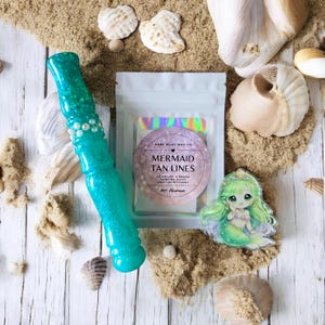 Puede incluir: Una varita turquesa brillante con detalles de perlas, una bolsa de ceras perfumadas Mermaid Tan Lines y una pegatina de sirena están dispuestas sobre arena con conchas marinas. El embalaje de la cera perfumada presenta el texto "Mermaid Tan Lines".