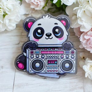May include: A sticker featuring a cartoon panda holding a retro boombox. The panda has black and white fur with pink accents on its ears and paws. The boombox is silver with pink and blue details.