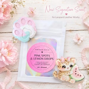 Può includere: Una bustina bianca con un'etichetta circolare rosa che riporta Pure Bliss Wax Co Pink Spots & Lemon Drops. Accanto si trovano una cera profumata a forma di zampa pastello e un leopardo in acrilico con un'anguria, circondati da fiori rosa.