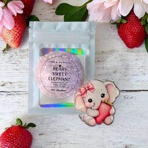 May include: A clear plastic bag containing a round, glittery wax melt with the text "Berry Sweet Elephant." A cartoon elephant holding a strawberry is next to the bag. Strawberries and pink flowers surround the bag.