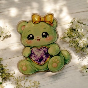 May include: A green teddy bear-shaped decoration with a yellow bow and a purple heart-shaped gem. The bear has large, expressive eyes and rosy cheeks. The design is detailed with paw prints on the feet.
