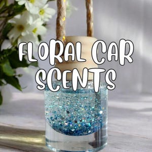 May include: A clear glass car air freshener with blue glitter and a wooden top. The text "FLORAL CAR SCENTS" is displayed in white. A natural rope is attached for hanging. White flowers are in the background.