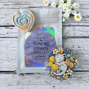 May include: A heart-shaped wax melt with blue, orange, and white layers sits next to a package with a circular label that reads "Sunfire Citrus." A decorative pin features a sleeping white dragon surrounded by citrus fruits and flowers.