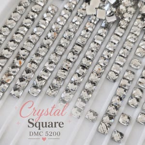 May include: A white plastic tray containing multiple rows of clear, square-cut faceted crystals arranged in vertical lines. The text Crystal Square DMC 5200 is printed in the lower left corner, accompanied by small decorative star and heart icons.