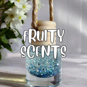 May include: A clear glass air freshener with blue glitter and a wooden top, suspended by a rope. The text "FRUITY SCENTS" is displayed in white. White flowers are visible in the background.