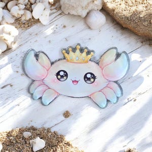 Può includere: Un adesivo a forma di granchio in stile cartone animato con una corona d'oro. Il granchio ha grandi occhi espressivi e una colorazione rosa e bianca. L'adesivo è su una superficie di legno bianco con sabbia e conchiglie.