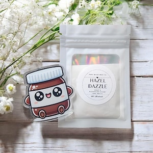 Puede incluir: Un colgante acrílico transparente con forma de tarro sonriente de crema de avellanas con tapa blanca. Junto a él, una bolsa blanca con ventana transparente, que muestra el texto "HAZEL DAZZLE".