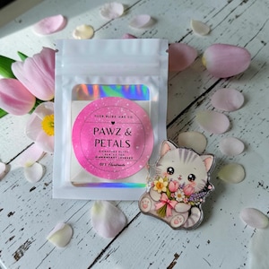 Op de afbeelding: Een doorzichtig zakje met een roze en wit label met de tekst "PAWZ & PETALS" en een katvormige speld met bloemen. Het label zegt ook "PURE BLISS WAX CO" en "100% Handmade". Roze en witte bloemblaadjes zijn verspreid.