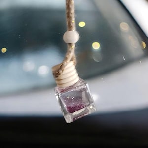May include: A small, clear glass bottle filled with purple glitter and a white bead on a brown string. The bottle is hanging from a rearview mirror.