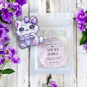 May include: A white cat sticker holding a diamond, a round sticker with "Sticky Pawz" text, and a clear bag. The stickers are on a white wooden surface, with purple flowers. The text on the round sticker is in English.