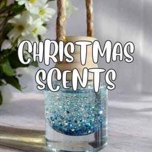 Könnte beinhalten: Ein klarer Glas-Lufterfrischer mit blauem Glitzer und dem Text "CHRISTMAS SCENTS". Der Lufterfrischer hat einen Holzaufsatz und ein Seil zum Aufhängen. Der Hintergrund umfasst weiße Blumen.