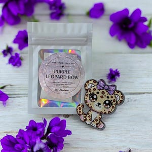 May include: A clear bag containing a round, glittery label with the text "PURPLE LEOPARD BOW" and a cute leopard-print bow-wearing animal. The label reads "LEOPARD LEATHER WORKS SIGNATURE PUTTY EDITION 2025 100% Handmade". Purple flowers are scattered around the objects.
