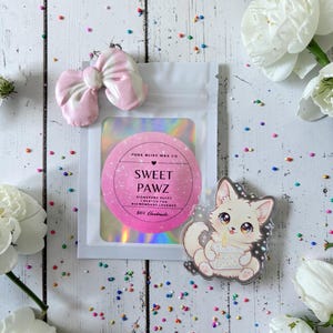 Op de afbeelding: Een platte lay-out met een roze en witte strik, een doorzichtige zak met een roze en wit Sweet Pawz label en een kattensticker met een taart. Het label luidt "Pure Bliss Wax Co" en "100% Handmade". De achtergrond is een wit houten oppervlak.