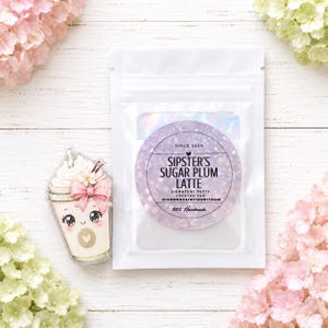 May include: A white resealable pouch containing putty with a circular label reading SIPSTER'S SUGAR PLUM LATTE SINCE 2024, accompanied by a clear acrylic charm shaped like a coffee cup with a pink bow, whipped cream, and a smiling face.