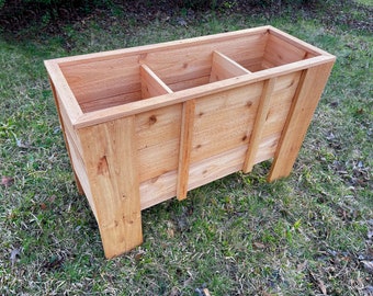 Op de afbeelding: Een houten plantenbak met drie compartimenten. De plantenbak is gemaakt van natuurlijk cederhout en heeft een lichtbruine afwerking. De plantenbak staat op een bed van groen gras.
