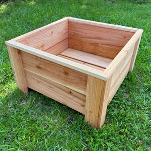 May include: A square wooden planter box made of light brown wood. The planter has a simple design with a rectangular shape and is sitting on a bed of green grass. The box is empty and ready for planting.