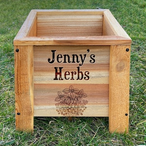 May include: A rectangular wooden planter box with the text "Jenny's Herbs" and a botanical illustration. The box is made of light-colored wood and has a natural finish. The planter is sitting on green grass.