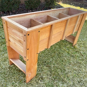 May include: A wooden raised garden bed with three compartments. The bed is made of natural wood and has a light brown finish. The bed is sitting on a grassy lawn.