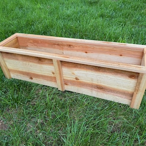 May include: A rectangular wooden planter box made of light brown wood, resting on green grass. The planter has a simple, open design with visible wood grain and knots. It is suitable for outdoor gardening.