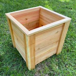 May include: A square wooden planter box made of light brown wood. The planter has a simple, modern design with a slightly tapered shape and is sitting on green grass. The box is constructed with visible wood grain.